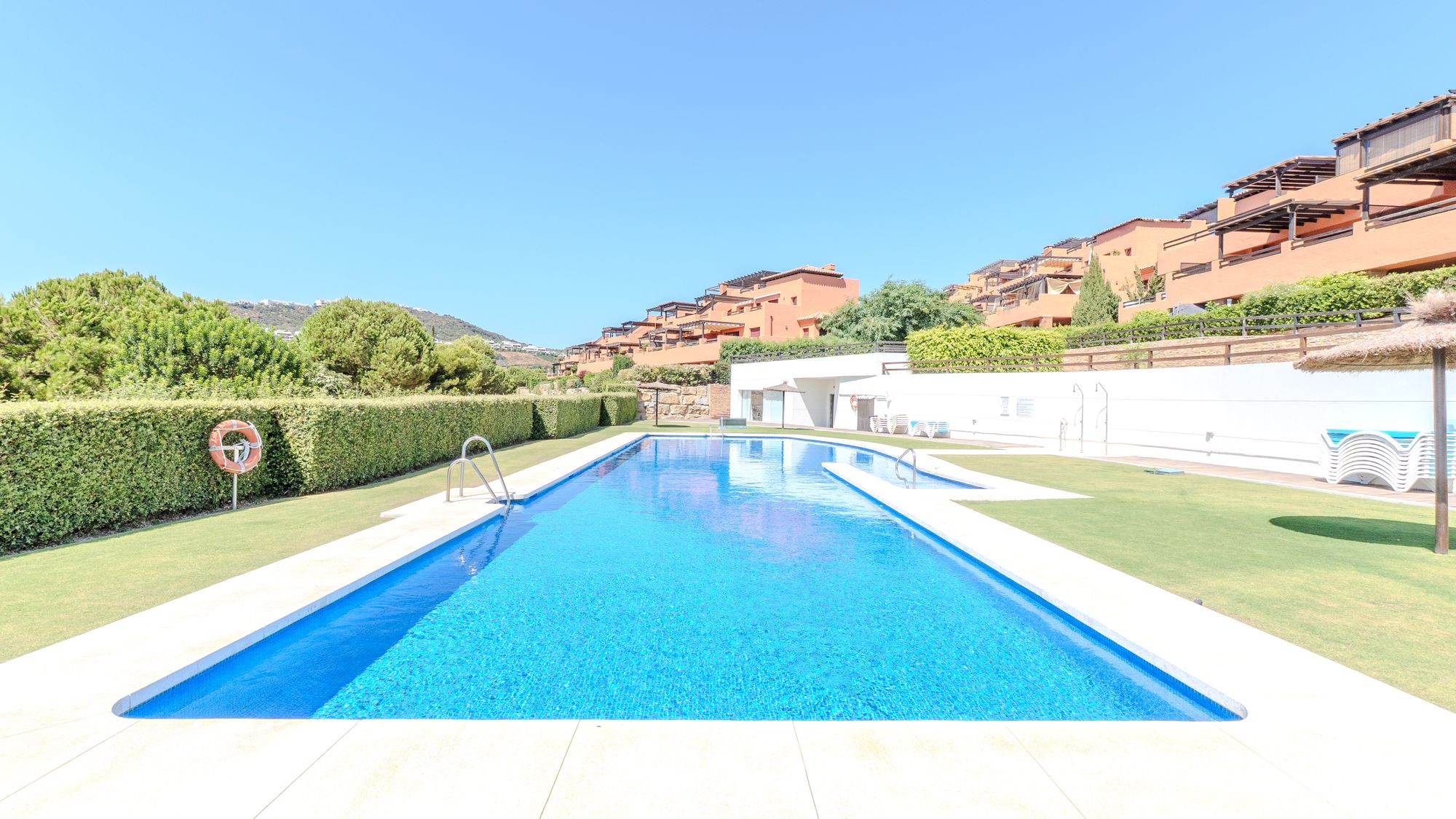 Long communal swimming pool surrounded by lawns, hedges and hillside homes